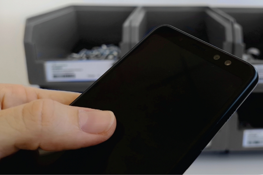 Eine Person hält ein Smartphone mit schwarzem Bildschirm in der Hand. Im Hintergrund sind mehrere graue Behälter mit kleinen technischen Produkten aus Metall zu sehen. Das Bild ist auf die Hand und das Telefon fokussiert, wobei die Behälter leicht unscharf sind.